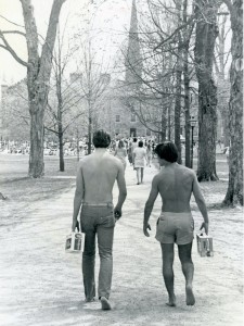 An archival photo from 1982 of the backs of two shirtless male students carrying six packs of beer and walking down Middle Path